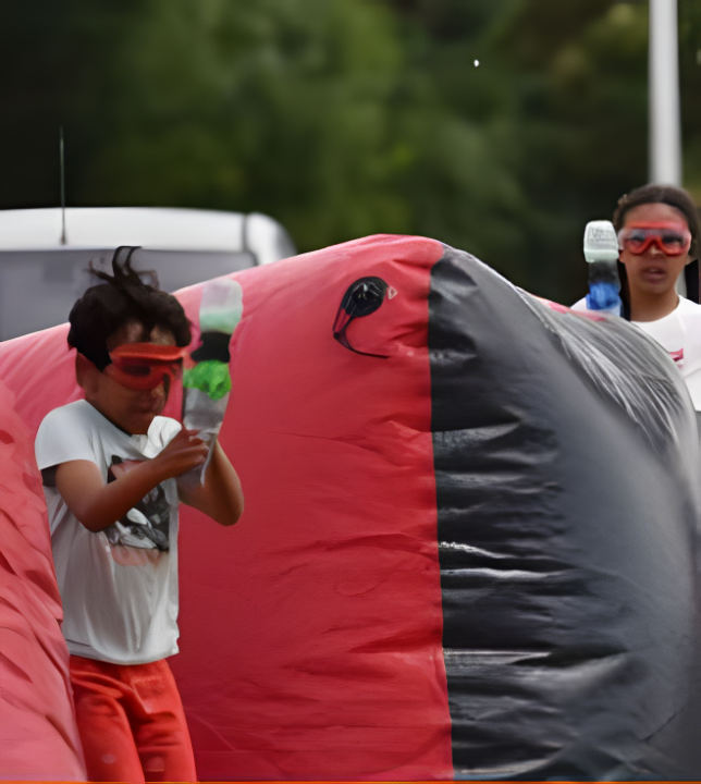 Un enfant en lunettes de protection, lanceur de mini ball en main, derrière un obstacle gonflable rouge
